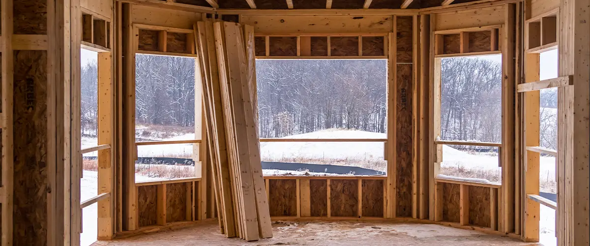 Framed home addition construction in winter showing exposed wood structure, large windows, and a scenic snowy landscape in the background.