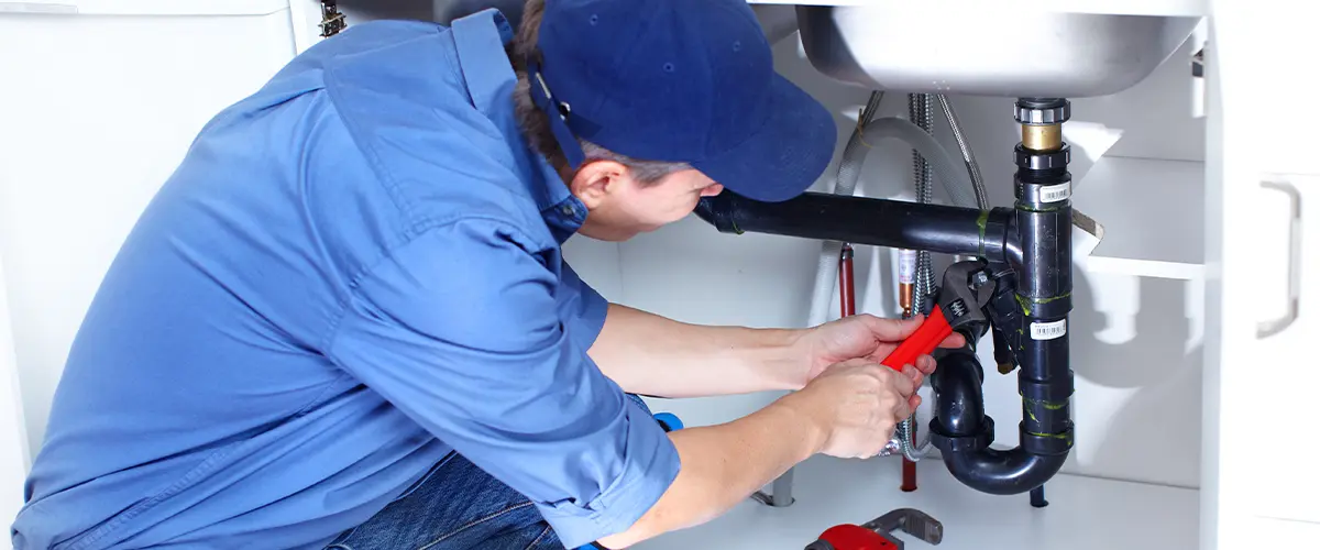 Professional plumber repairing kitchen sink plumbing with a wrench, wearing a blue cap and shirt, surrounded by tools.