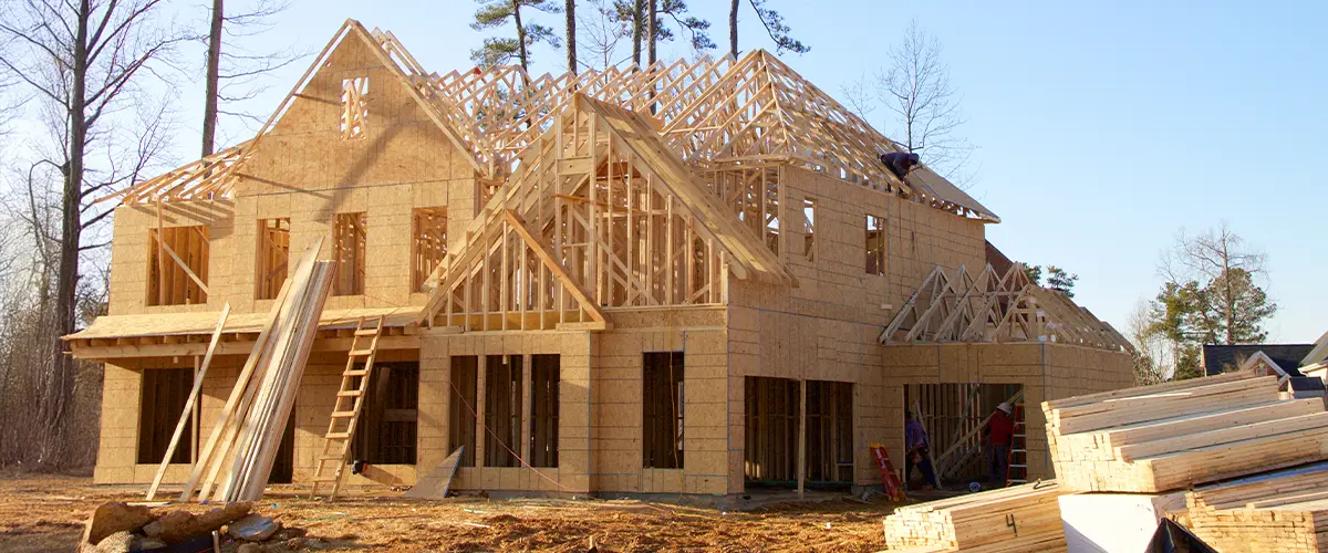 New home construction with wooden framing in progress, featuring roof trusses and exterior sheathing under a clear blue sky.